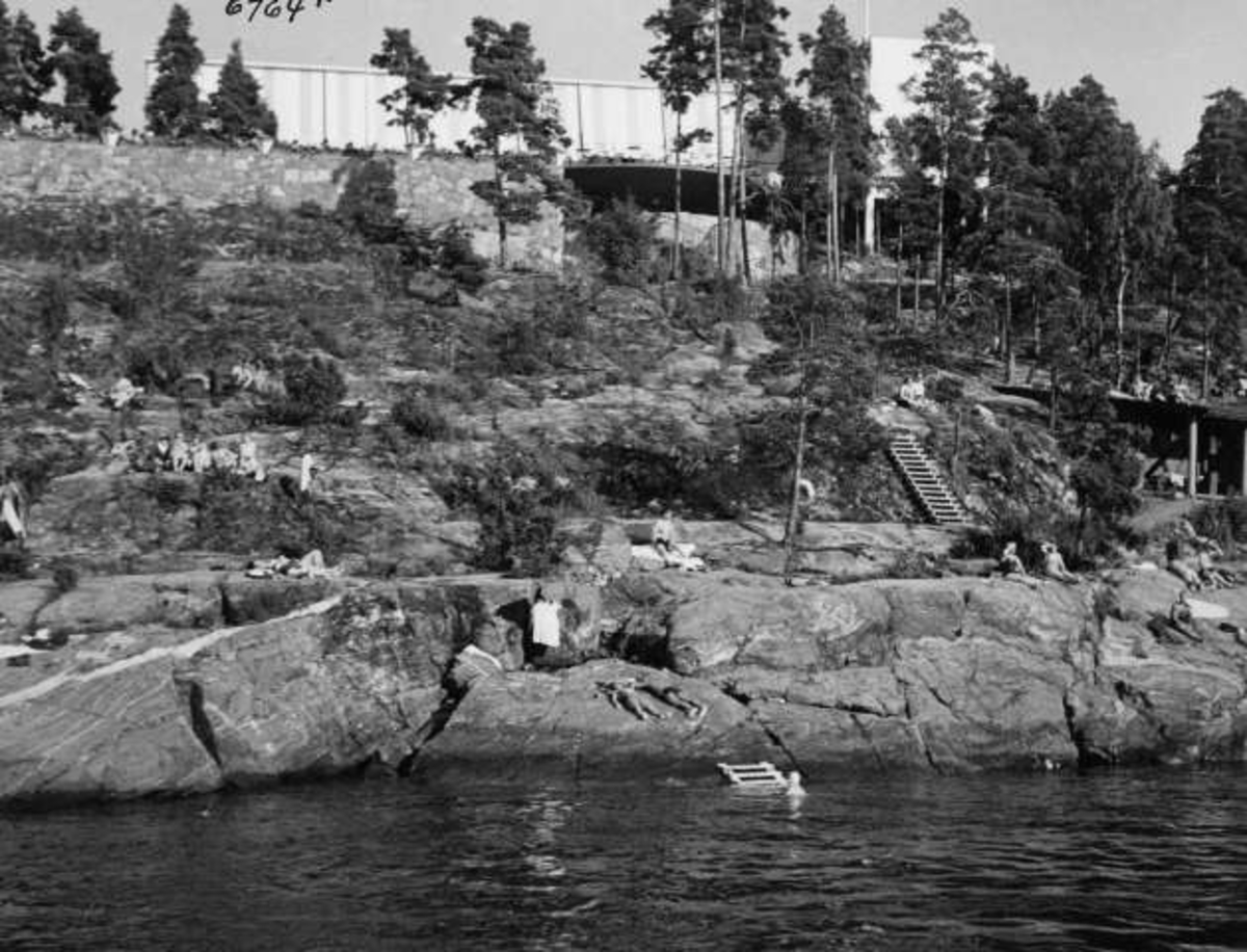 Anders Beer Wilse, Strandliv med badegjester som soler seg på svaberget ved sjøen på Ingierstrand bad utenfor Oslo. Restauranten i bakgrunnen.