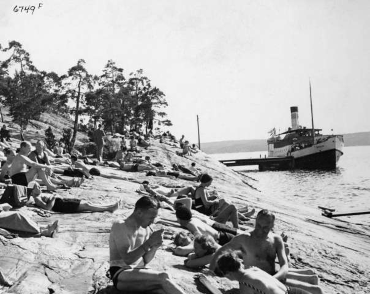 Anders Beer Wilse, Strandliv med badegjester som soler seg på svaberget ved sjøen på Ingierstrand bad utenfor Oslo. Båt ved brygga i bakgrunnen.