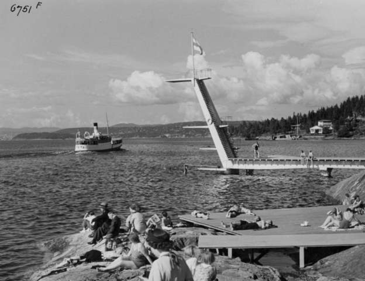 Anders Beer Wilse, Strandliv med badegjester som soler seg på svaberget ved sjøen på Ingierstrand bad utenfor Oslo. Stupetårn og båt i bakgrunnen.