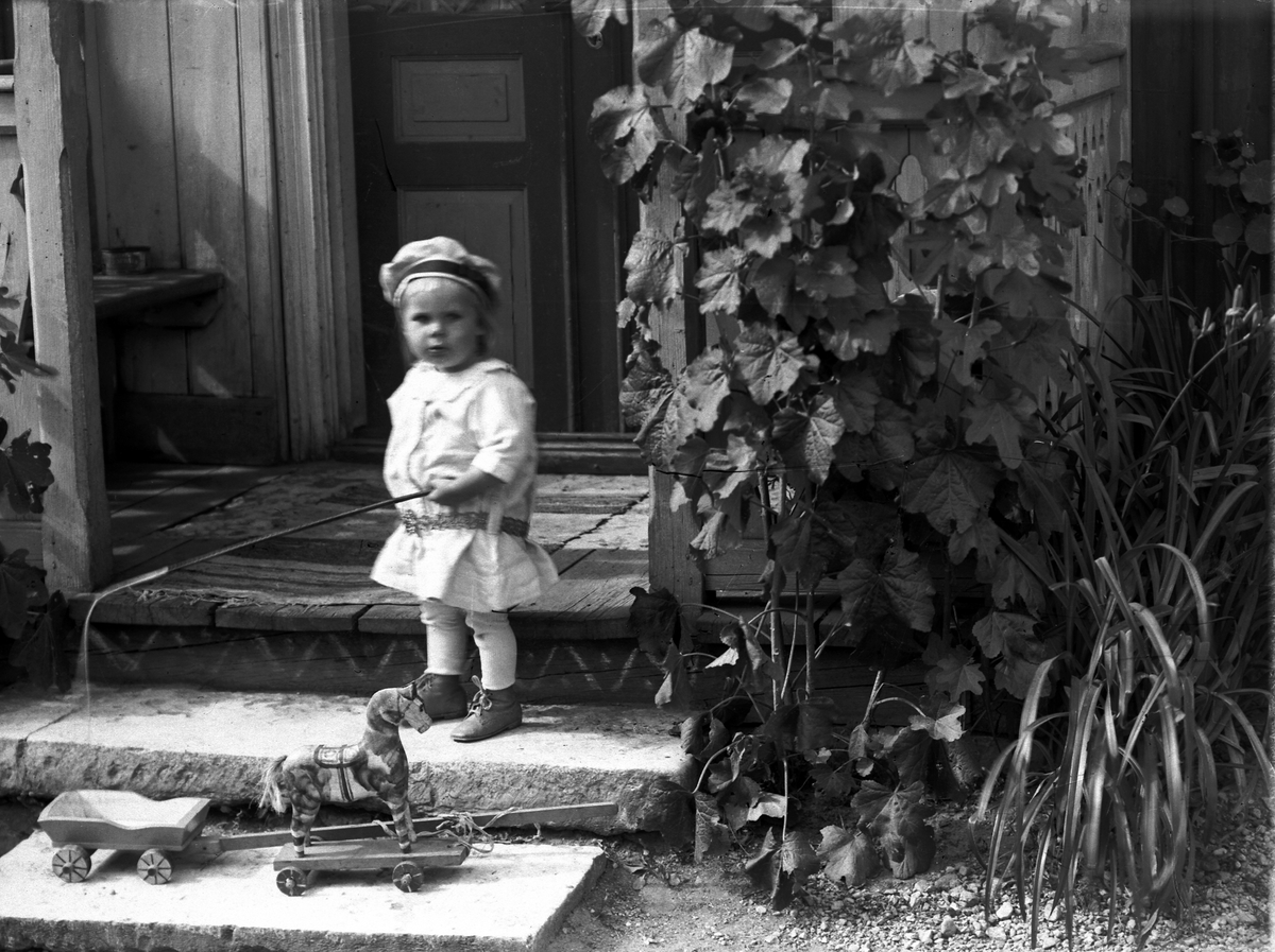 Samuel Lindskog, A young child with a toy horse and a carriage on the steps of the house. Oskar Nilsson