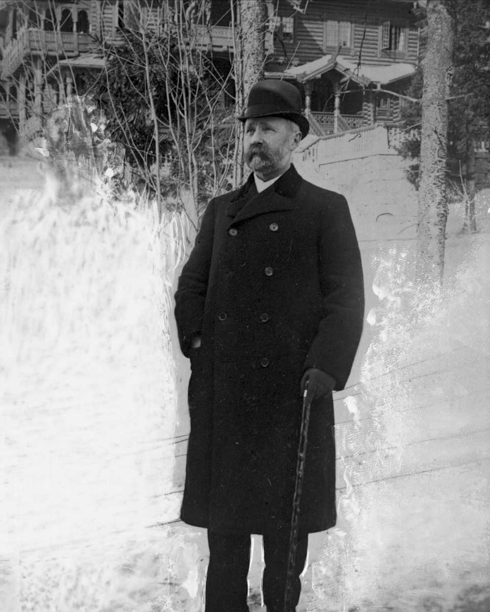 Gustav Borgen, Avfotografering. Portrett, mann utendørs vinterstid, med hatt, frakk og stokk. Bygninger i bakgrunnen.