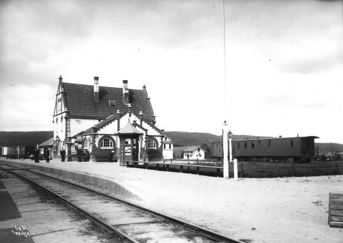 Anders Beer Wilse, Sørumsand stasjon, Sørum, Akershus 1907. Stasjonen, reisende på plattformen, kiosk og jernbanevogn.