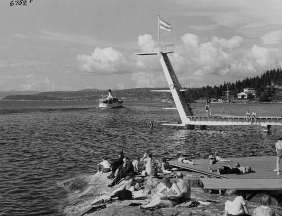 Anders Beer Wilse, Strandliv med badegjester som soler seg på svaberget ved sjøen på Ingierstrand bad utenfor Oslo. Stupetårn og båt i bakgrunnen.