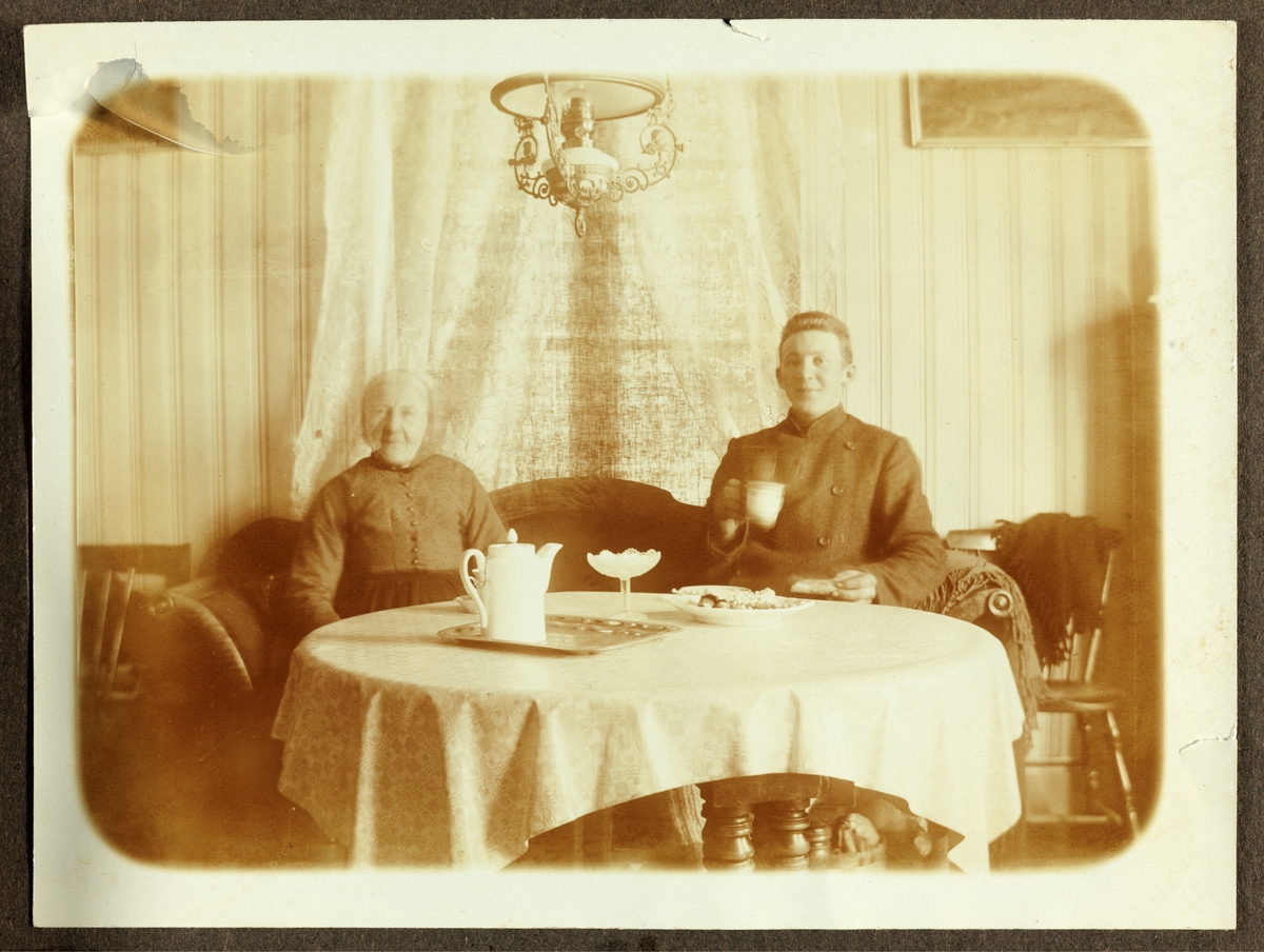 Unknown artist, Interiørbilde fra gartnerboligen på Bygdø Kongsgård. To ukjente personer ved kaffebord med kaker.