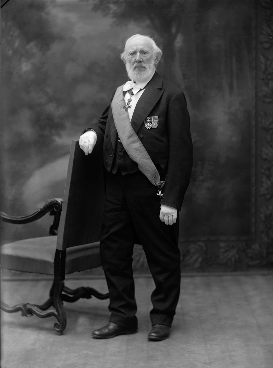 Gustav Borgen, Portrett, S.H. Lundh (Sverrer Hakon), gift 1867 med Sophie f. Warmuth. Han var forretningsmann, maskin- og konsulentforretning. Mann i dress med medaljer og ordener poserer ved høyrygget stol.