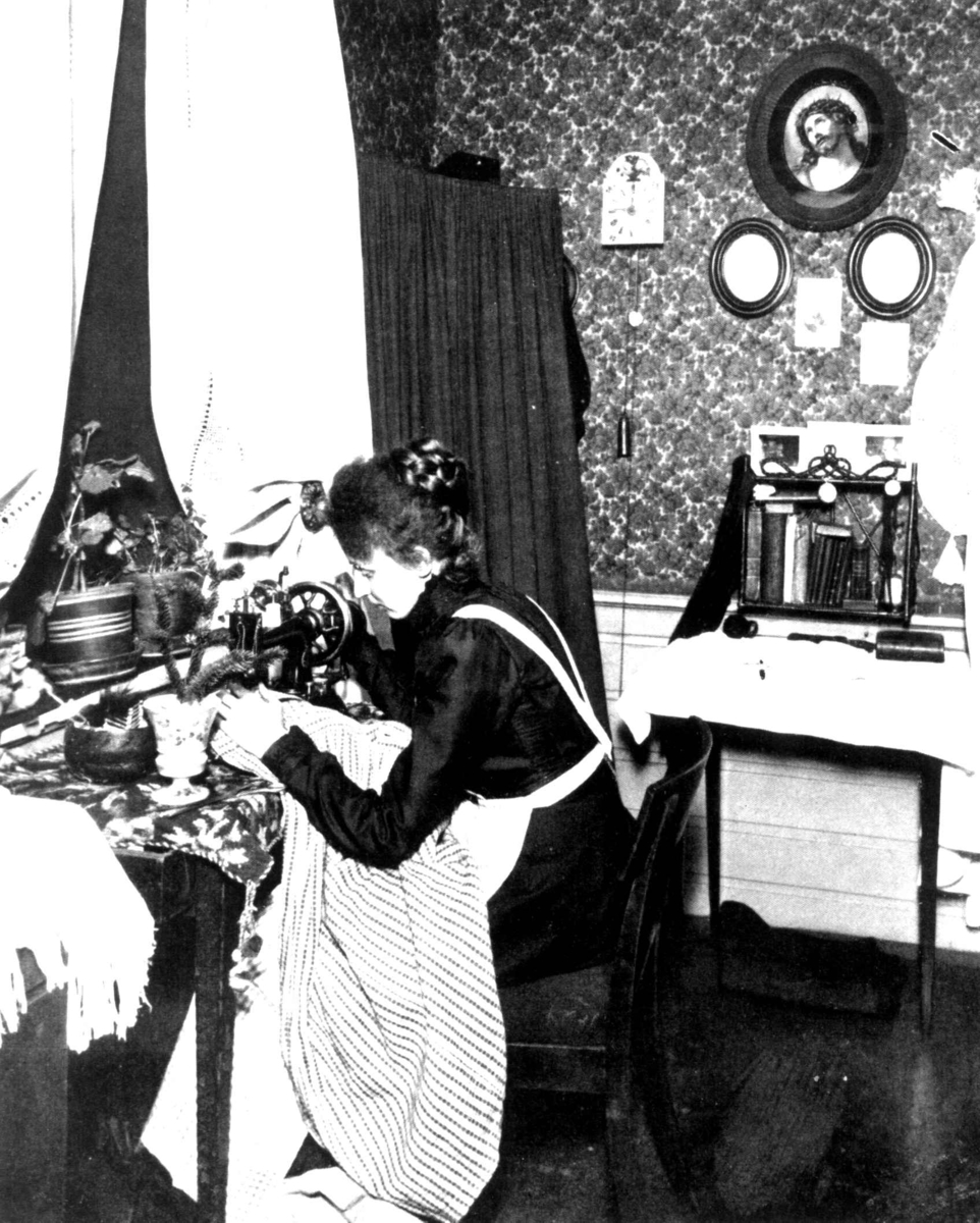 Anders Beer Wilse, Sypike, "Sypikens julaften". 1902. En kvinne sitter i et rom og syr på symaskin.