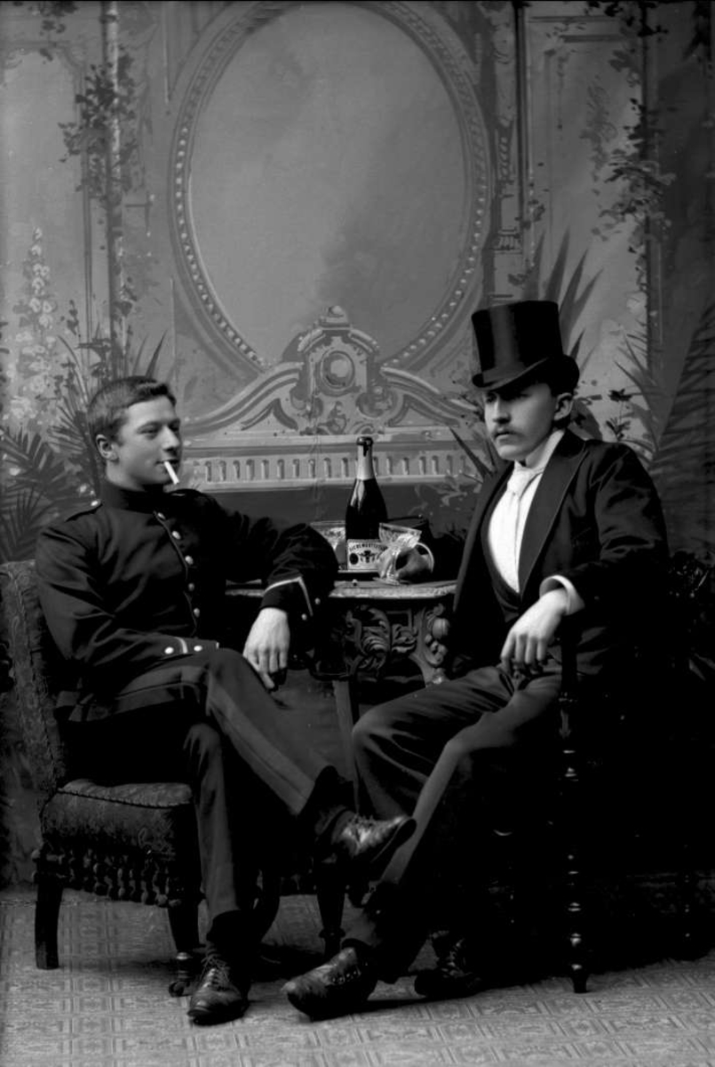 Gustav Borgen, Gruppeportrett, to menn sittende ved bord, champagne og sigarett.
