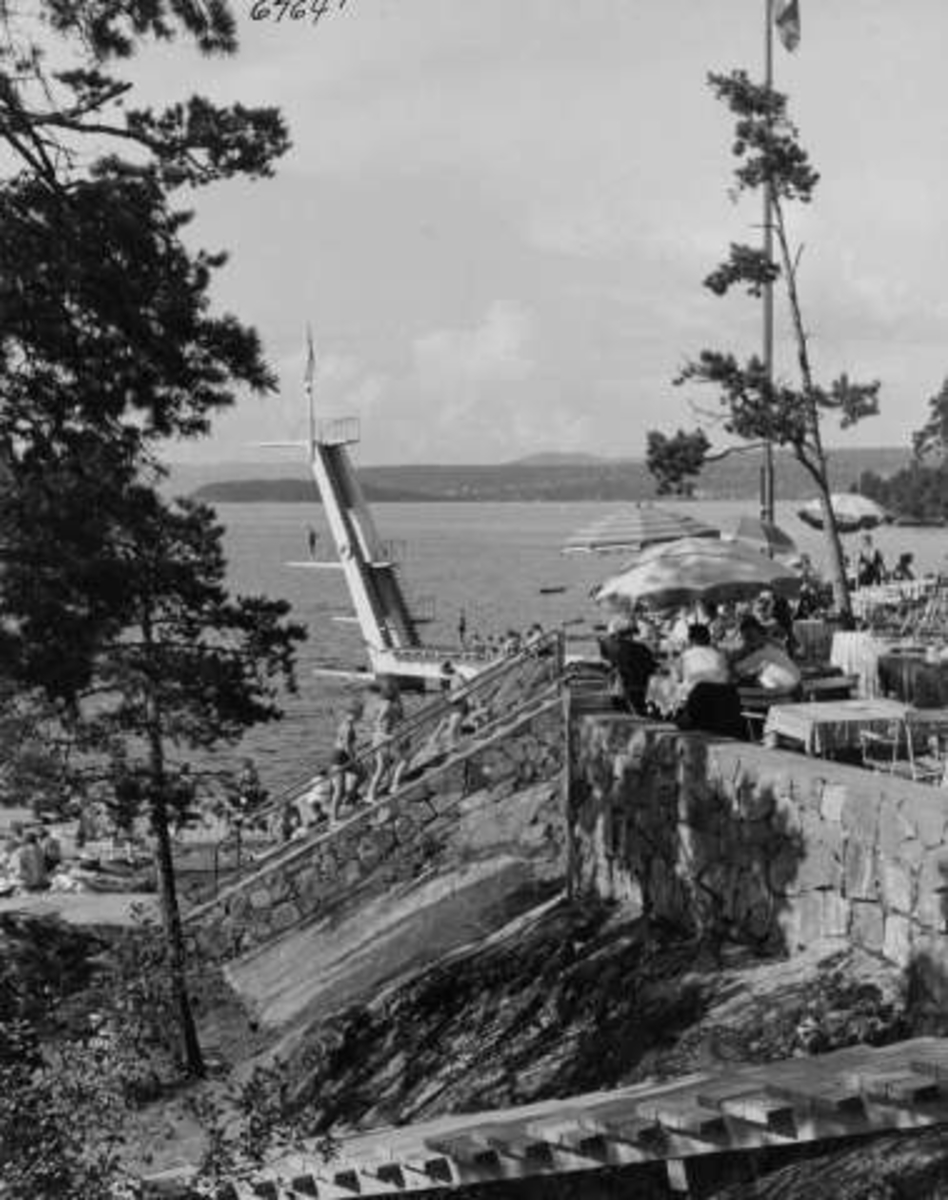 Anders Beer Wilse, Strandliv med badegjester ved sjøen på Ingierstrand bad utenfor Oslo. Stupetårn i bakgrunnen for terrassen med kafé og gjester under parasoller.