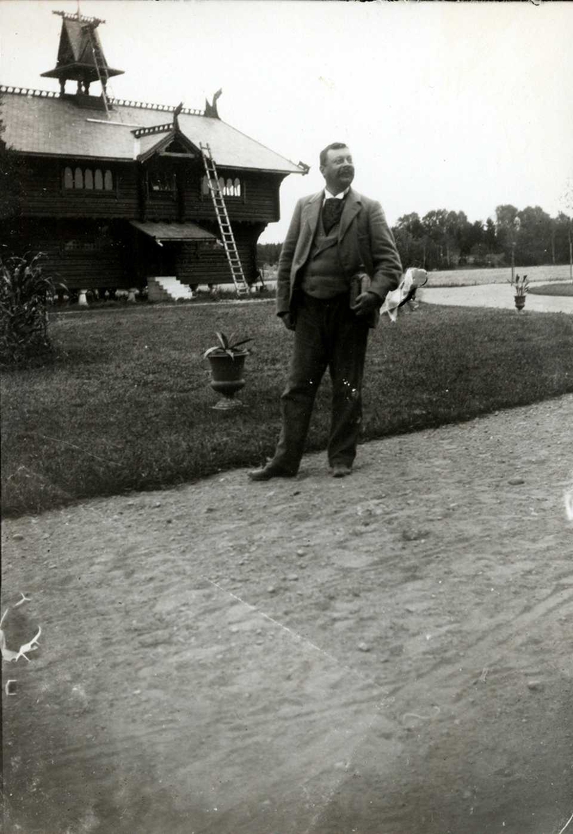 Fredrik Emil Faye, Portrett, ukjent mann, stående utendørs på tun foran trebygning i dragestil, muligens Evje herregård, Dilling, Rygge (jfr. NF.05269-151). Fra portrettserie av personer som bodde på eller besøkte Dal gård, Ullensaker, fotografert av gårdens eier, kammerherre Fred. Emil Faye (1844-1903) i årene 1875-…